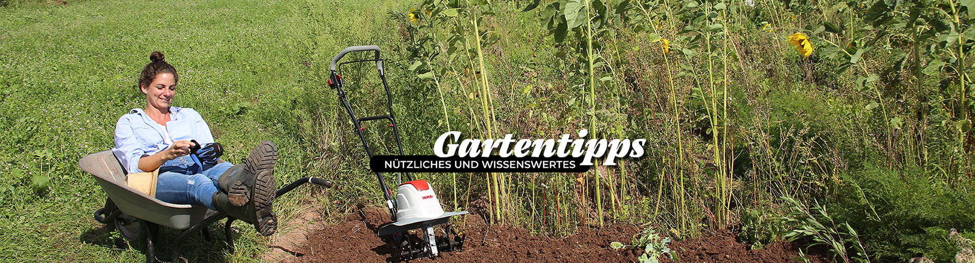 Frau sitzt lächelnd in einer Schubkarre in einem Beet mit Sonnenblumen neben ihr steht eine Ikra Bodenhacke